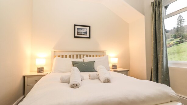 A neatly made bed with white linens and two rolled towels, a framed picture on the wall, and a window with curtains. - Waterhead Cottage (Ambleside)