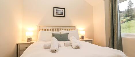 A neatly made bed with white linens and two rolled towels, a framed picture on the wall, and a window with curtains.