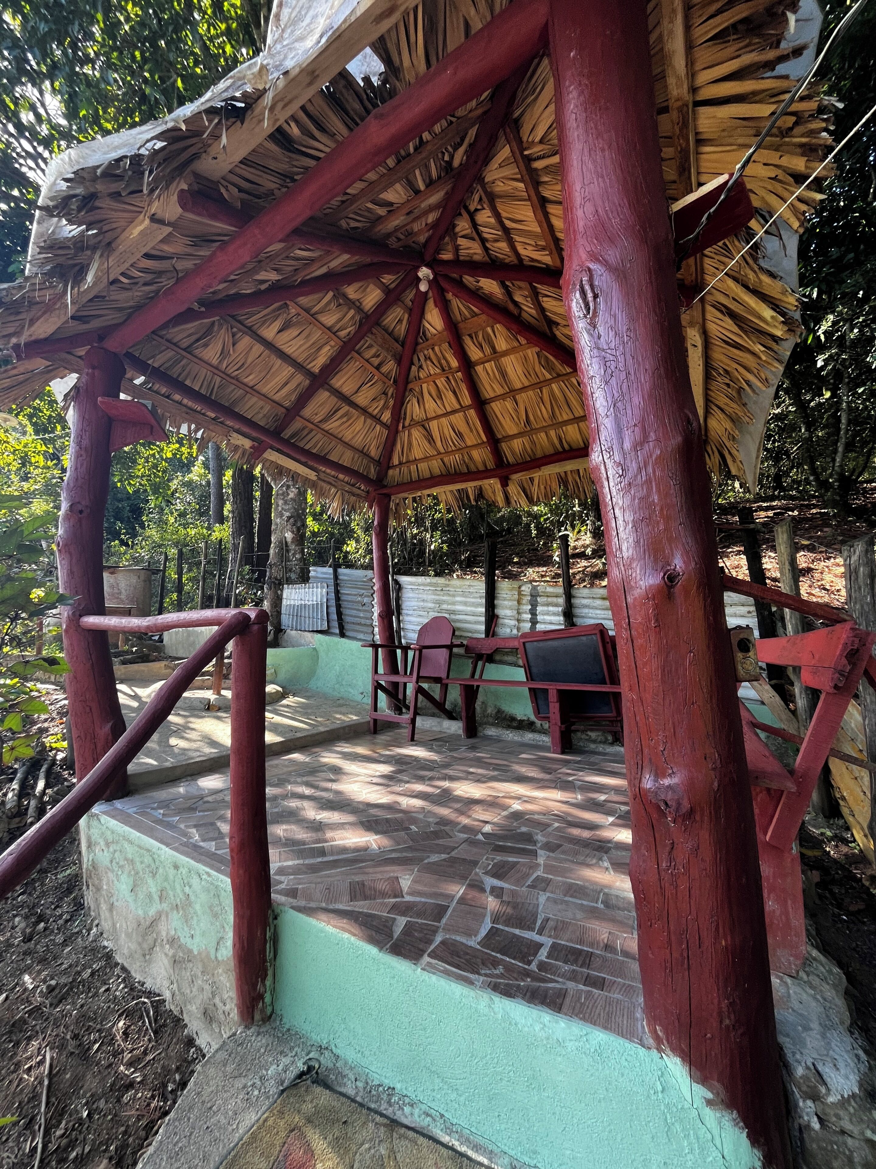 A wooden gazebo with a thatched roof, stone flooring, and red metal railings.