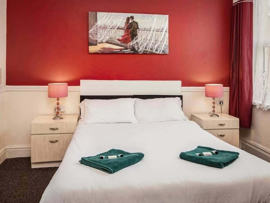 Quarto de hotel com parede de destaque vermelha, cama branca, duas mesas de cabeceira com abajures e um quadro sobre a cama.