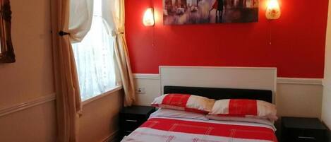 A hotel room with a red wall, a bed with a red and white bedspread, a painting above the bed, and two bedside lamps.