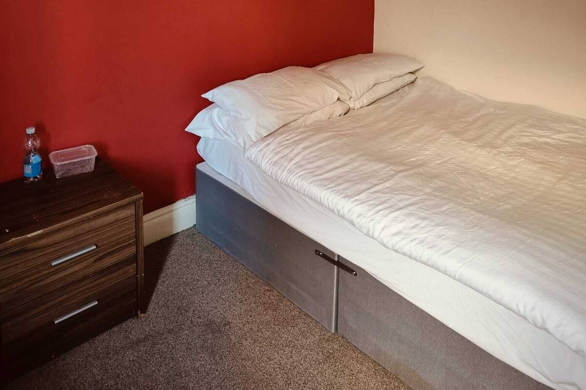 Ein Schlafzimmer mit einem Bett, einem Schminktisch, einer Wasserflasche, einem Plastikbehälter und einem Bild an der Wand.