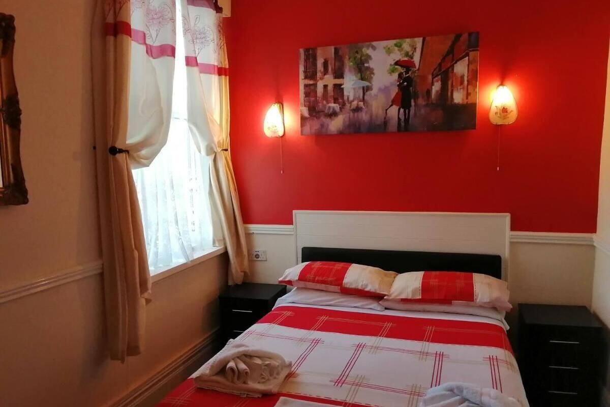Ein Hotelzimmer mit roter Wand, einem Bett mit rot-weiß kariertem Überwurf, zwei Nachttischlampen, einem Bild über dem Bett und einem Fenster mit Vorhängen.