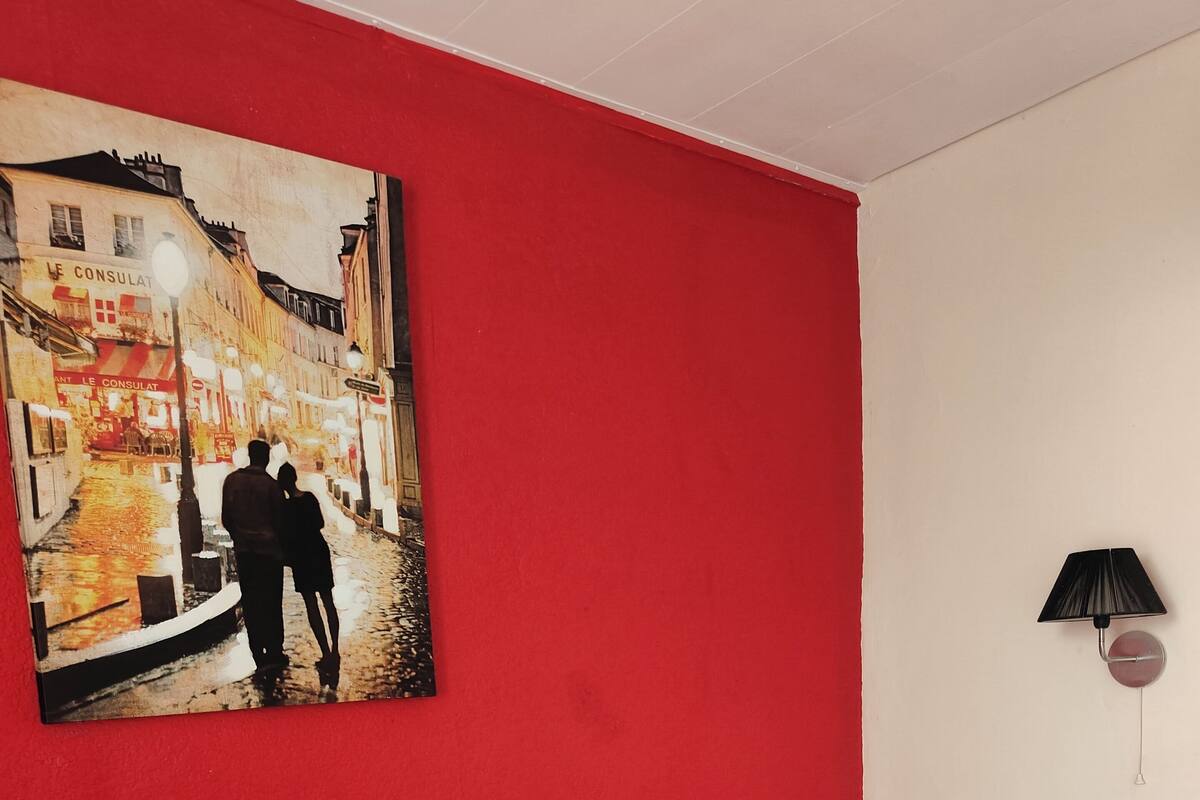 Ein Zimmer mit einem Einzelbett, einer roten Wand und einem Gemälde, das ein Paar auf einer Straße zeigt.