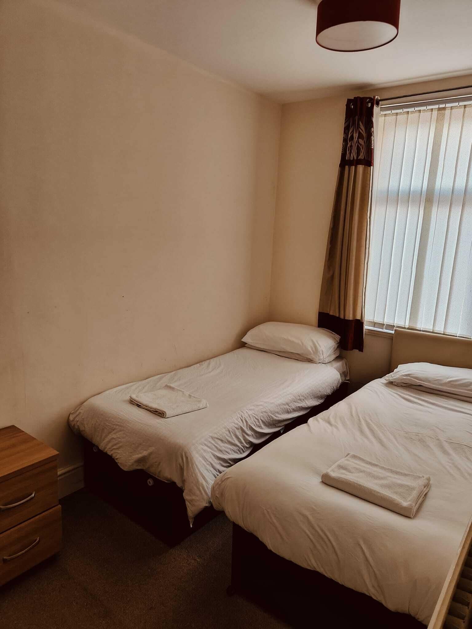 Een kleine, eenvoudige kamer met twee éénpersoonsbedden, een houten nachtkastje en een raam met gordijnen.