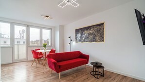 A modern living room with a red sofa, wooden flooring, a dining area with red chairs, and a large window offering a city view. - Apartament Grace Q4 Apartments (Gdynia)