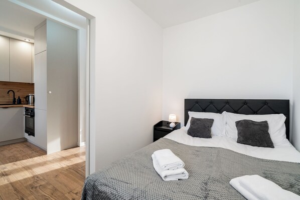 A modern bedroom with a neatly made bed, a nightstand with a lamp, and a view of a kitchen area. - Apartament Grace Q4 Apartments (Gdynia)