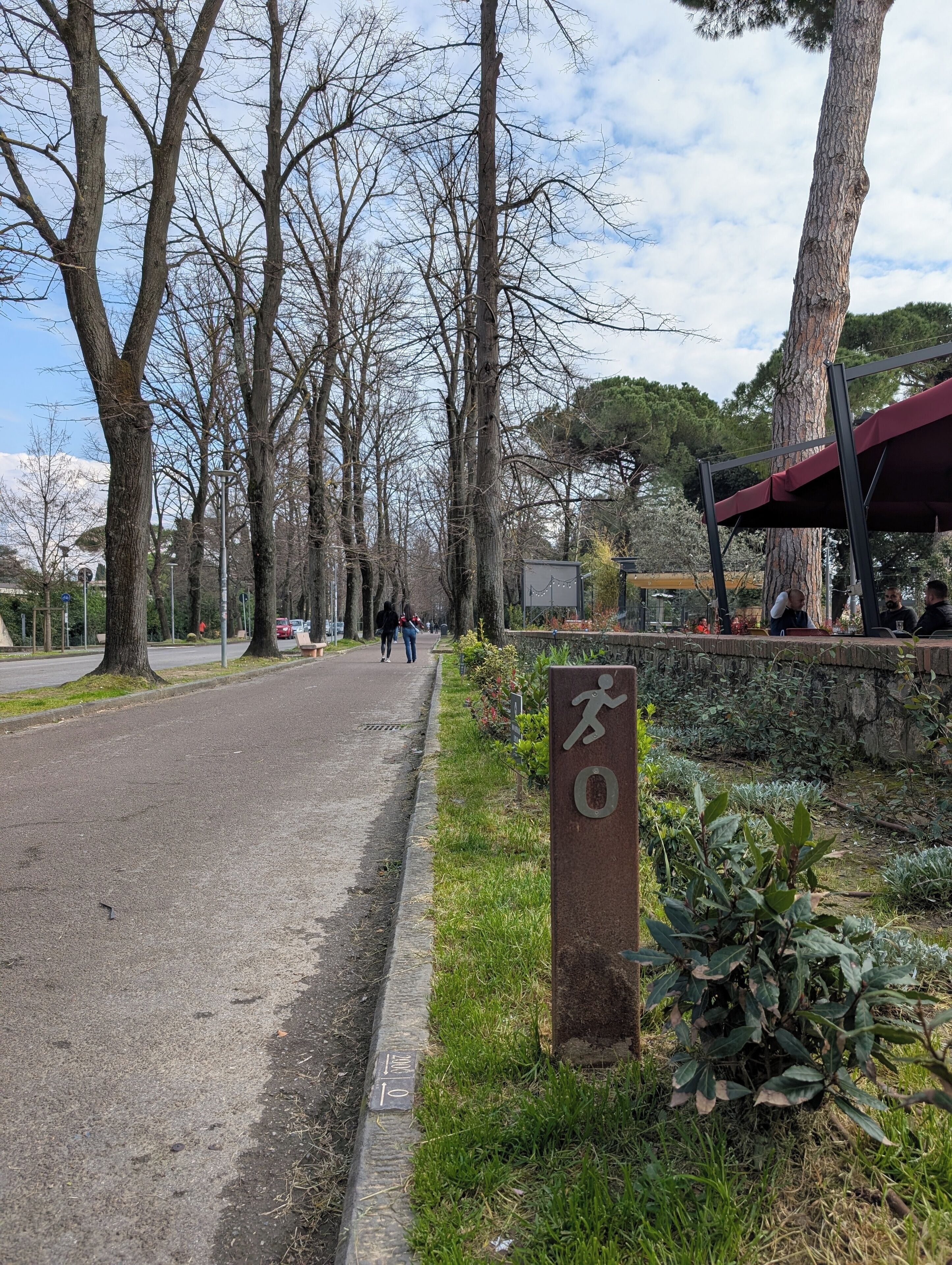 Un chemin pavé bordé d’arbres, un panneau représentant une personne en train de courir et un mur de pierres recouvert de verdure.