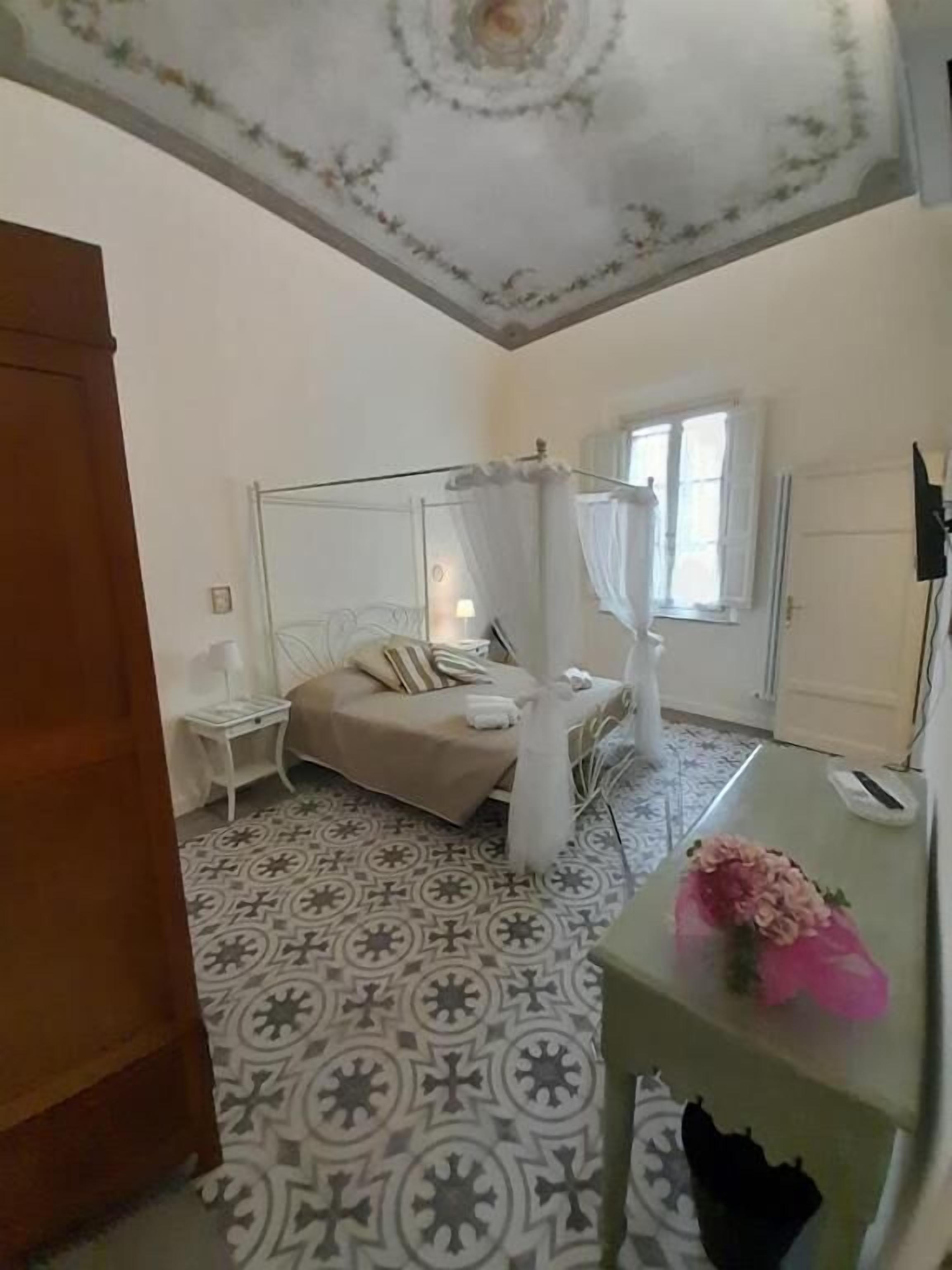 A bedroom with a canopy bed, a patterned floor, a small bedside table, and a desk with a flower.