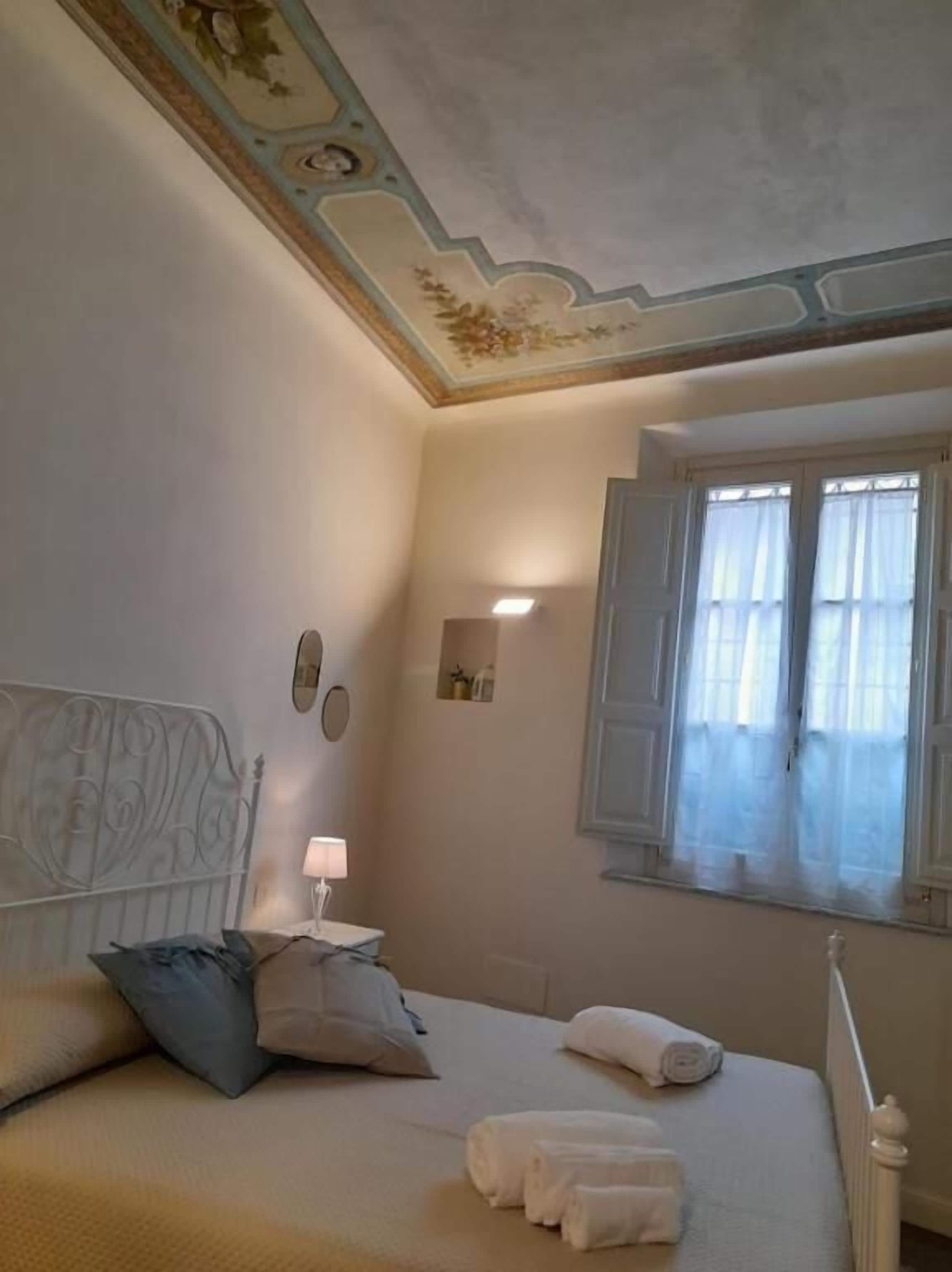 Une chambre à coucher comprenant un lit, des oreillers, une table de chevet avec une lampe, une fenêtre avec des rideaux et un plafond décoratif.