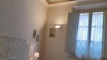 Une chambre à coucher comprenant un lit, des oreillers, une table de chevet avec une lampe, une fenêtre avec des rideaux et un plafond décoratif.