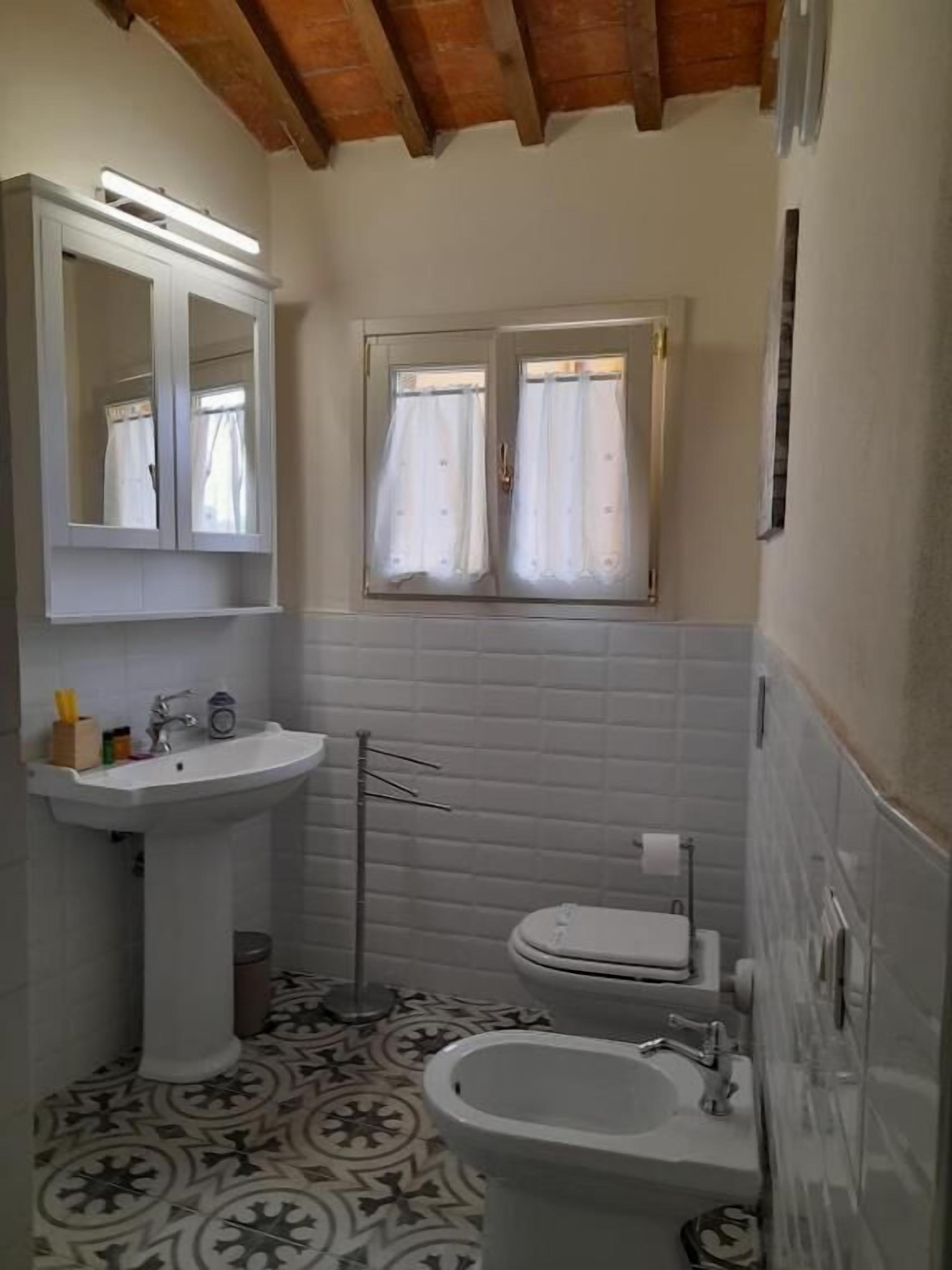 A small bathroom with a pedestal sink, a toilet, and a bidet. There is a window with curtains and a mirror above the sink.