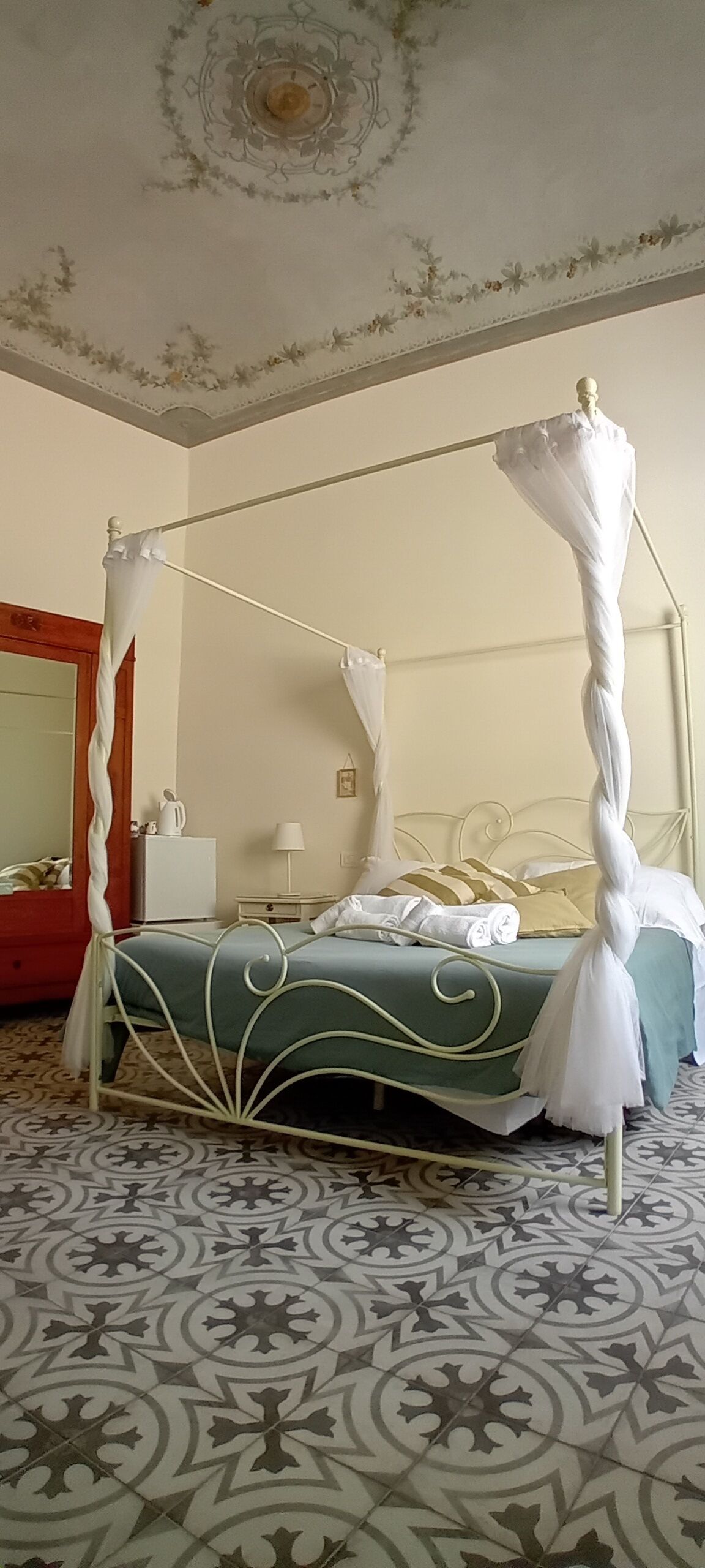 Ein Himmelsbett mit Metallgestell und weißem Baldachin, ein Nachttisch mit Lampe und ein dekorativer Spiegel an der Wand.