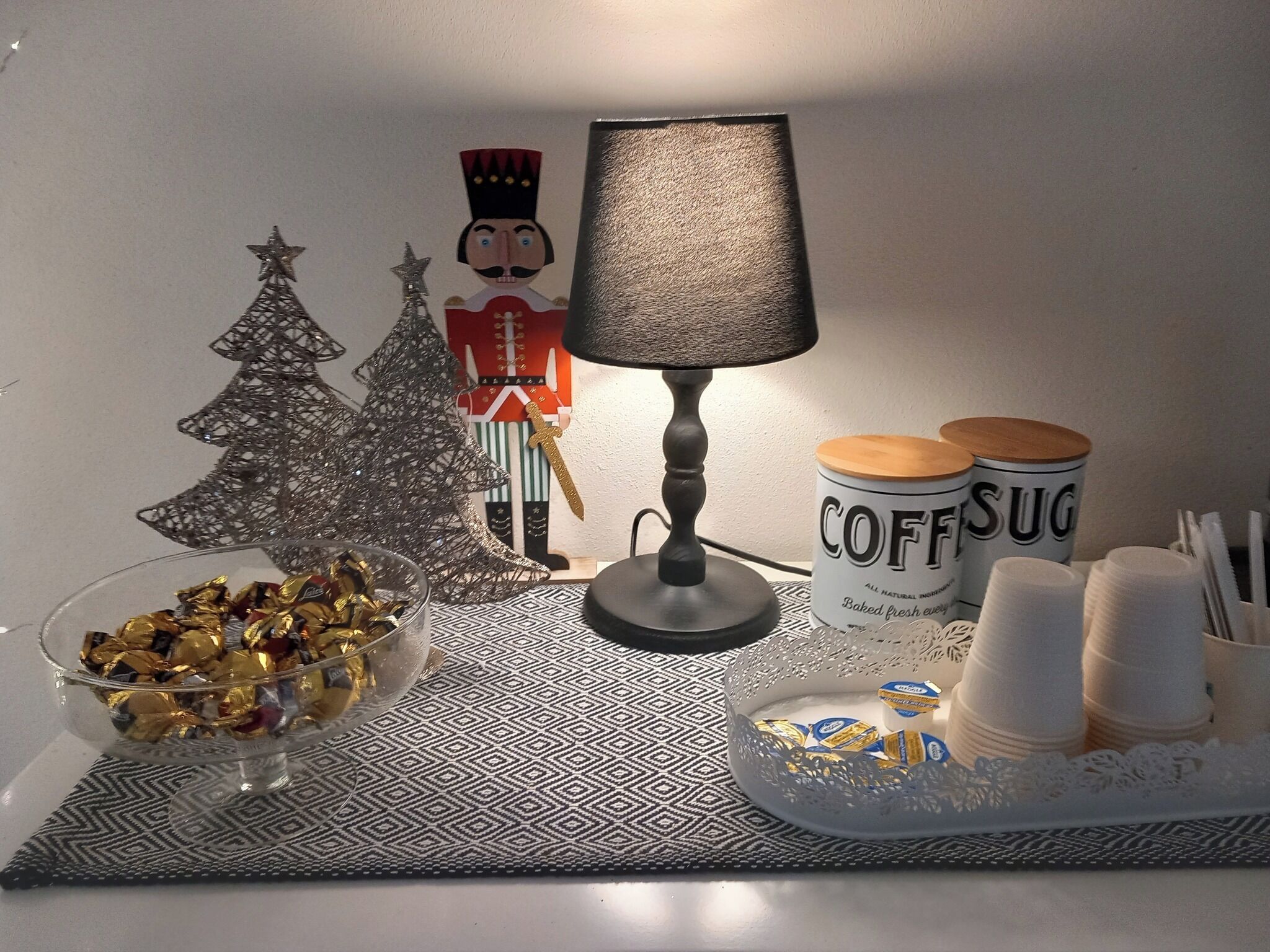 Una mesa con un cascabel decorativo, dos árboles de Navidad, una lámpara y una bandeja con tazas de café y un bol de bombones.