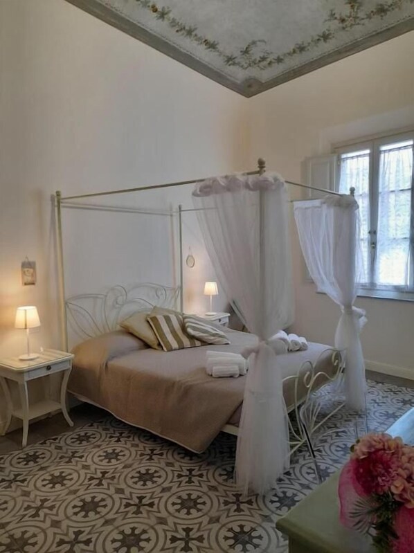 A four-poster bed with a canopy, two bedside tables with lamps, a patterned floor, and a window with curtains. - B&B Villino Luschi Pisa Centro (Pisa)