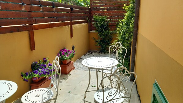 A small outdoor patio with a wooden pergola, white metal chairs, a round table, and potted plants.