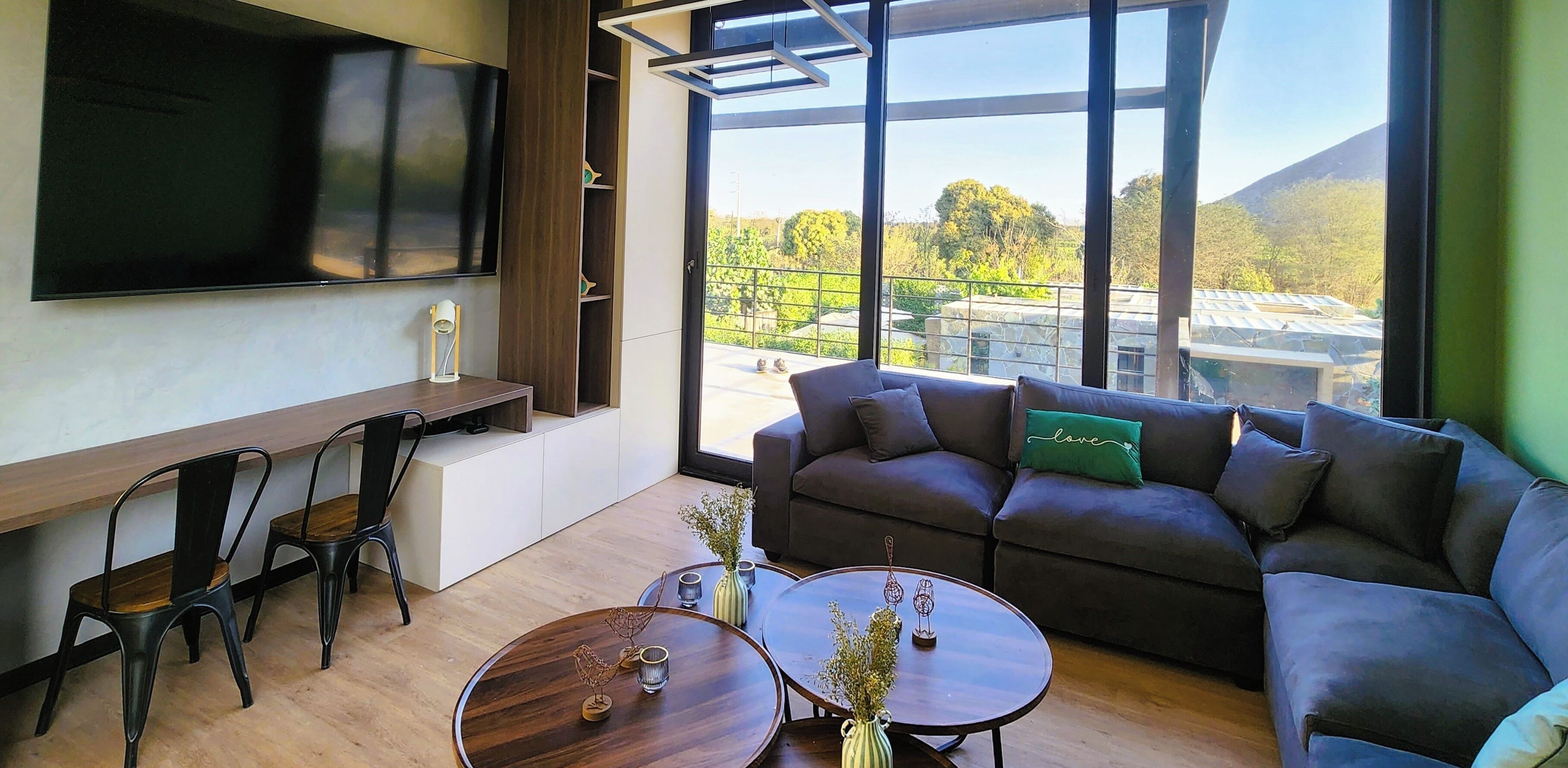 Un salón moderno con un televisor de pantalla plana grande, un sofá, dos mesas auxiliares y vistas a un balcón con vegetación y un edificio.