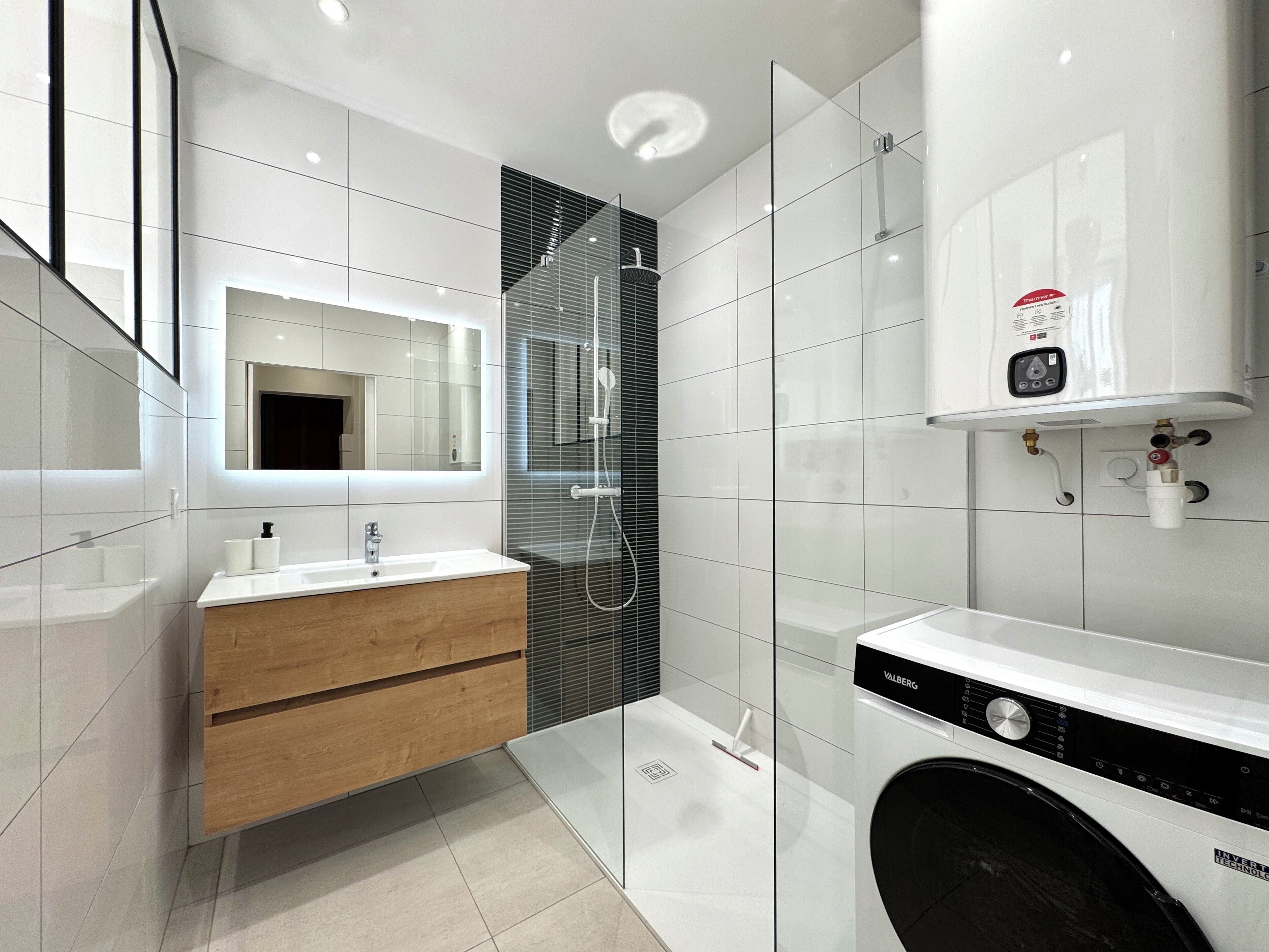Un baño moderno con cabina de ducha de vidrio, lavamanos integrado en un mueble de madera y lavadora empotrada.
