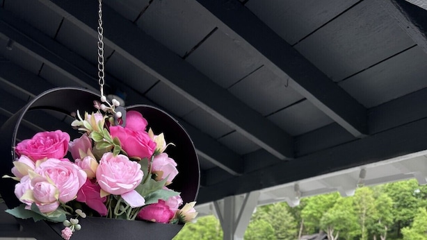 Un'area esterna riparata con un'elegante composizione floreale sospesa, un tavolo con un vaso rosa e vista su un lago con una casa sullo sfondo.