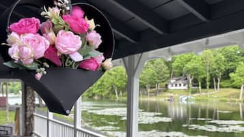 Un espacio al aire libre cubierto con un arreglo floral colgante, una mesa con un florero rosa, y una vista a un lago con una casa al fondo.
