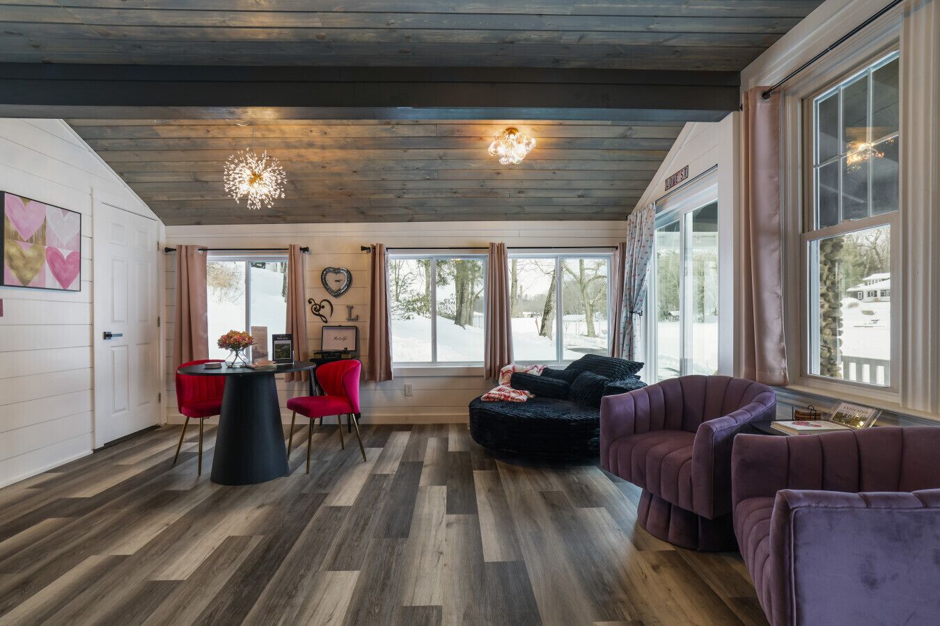 Una habitación acogedora con techo de madera, un sofá negro, una silla roja y un sillón morado. Amplias ventanas con cortinas, suelo de madera y una lámpara de pared.