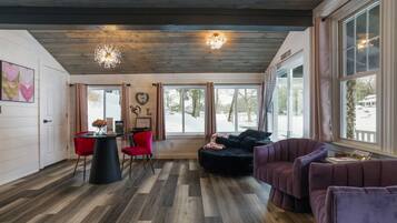 Una habitación acogedora con techo de madera, un sofá negro, una silla roja y un sillón morado. Amplias ventanas con cortinas, suelo de madera y una lámpara de pared.