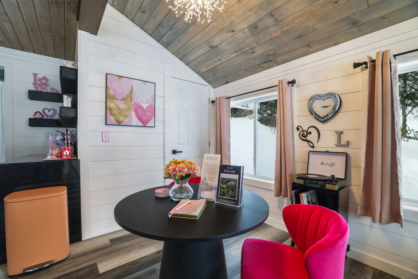 Una habitación moderna con una mesa redonda, una silla roja, un florero con flores, un cartel de bienvenida y una pared con un adorno con forma de corazón.