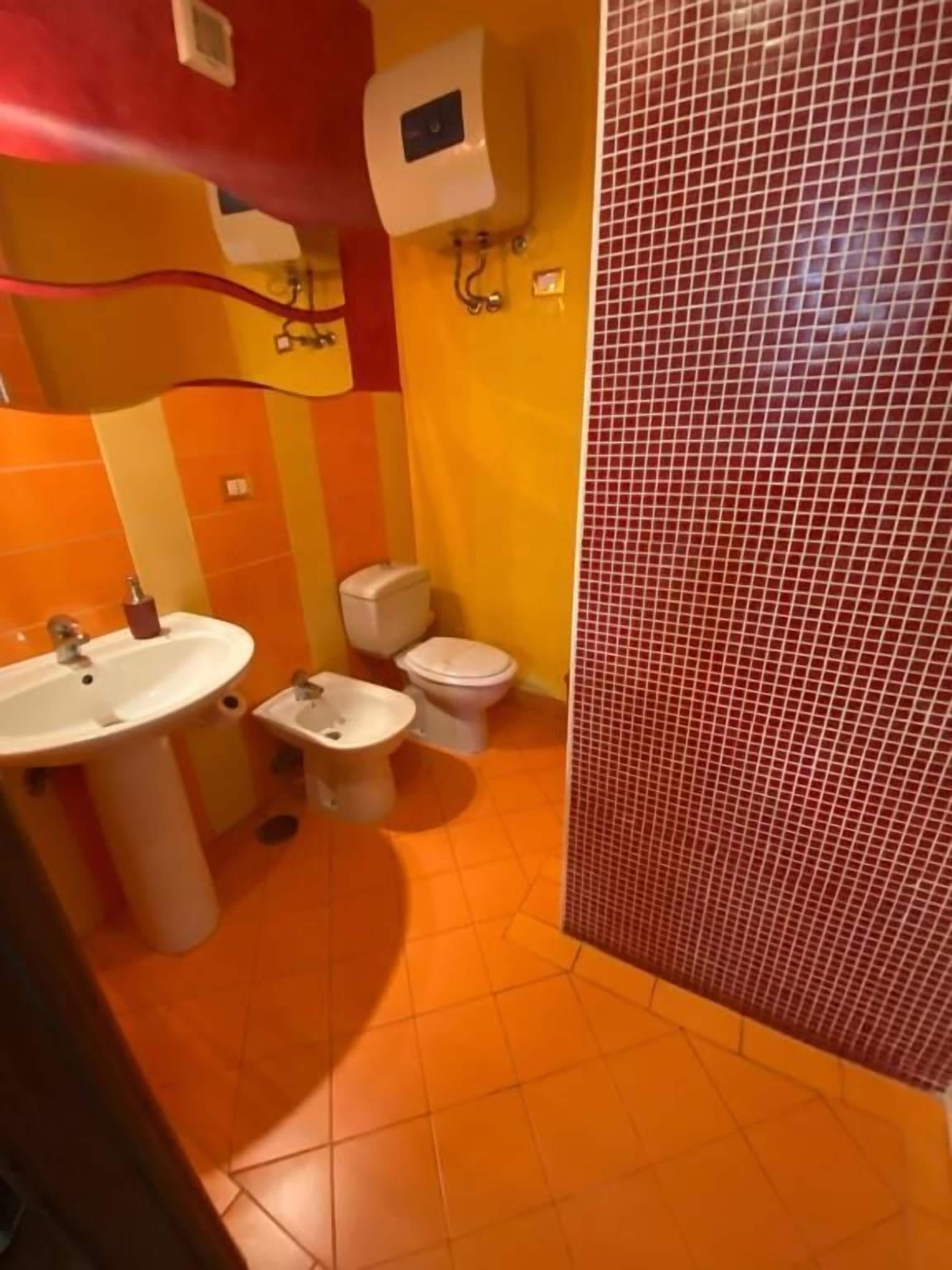 A bathroom with a sink, toilet, and bidet. The walls are tiled in orange and red, and there is a red door.