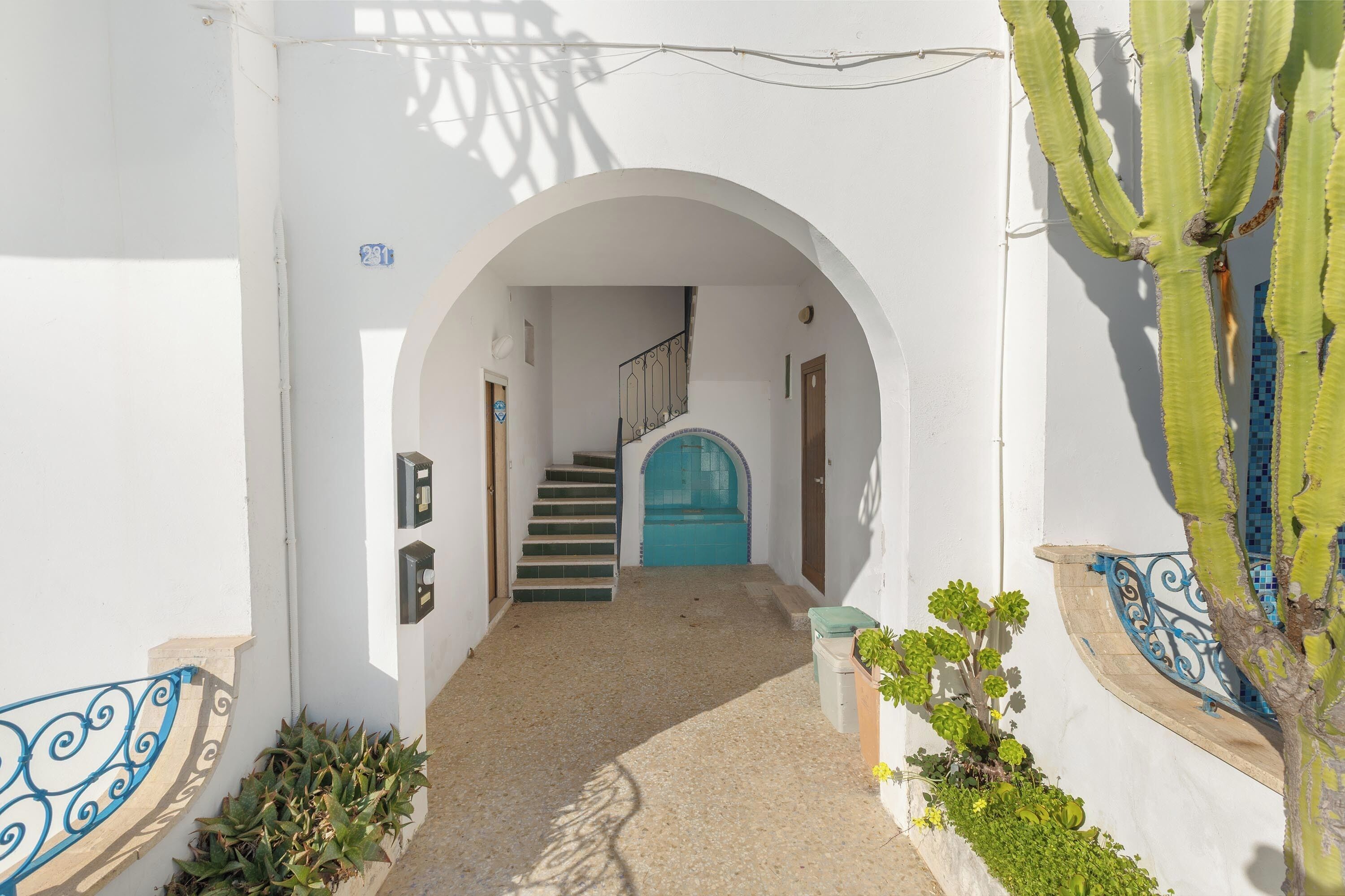 Un étroit sentier mène à un escalier orné d’une porte bleue, le tout étant bordé de murs blancs et de rampes de protection en métal décoratives.
