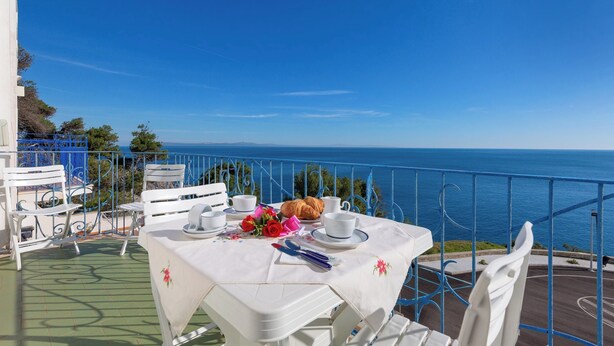 Et bord dækket til to personer med en croissant, en buket røde roser og en blå pen på en balkon med udsigt over havet.