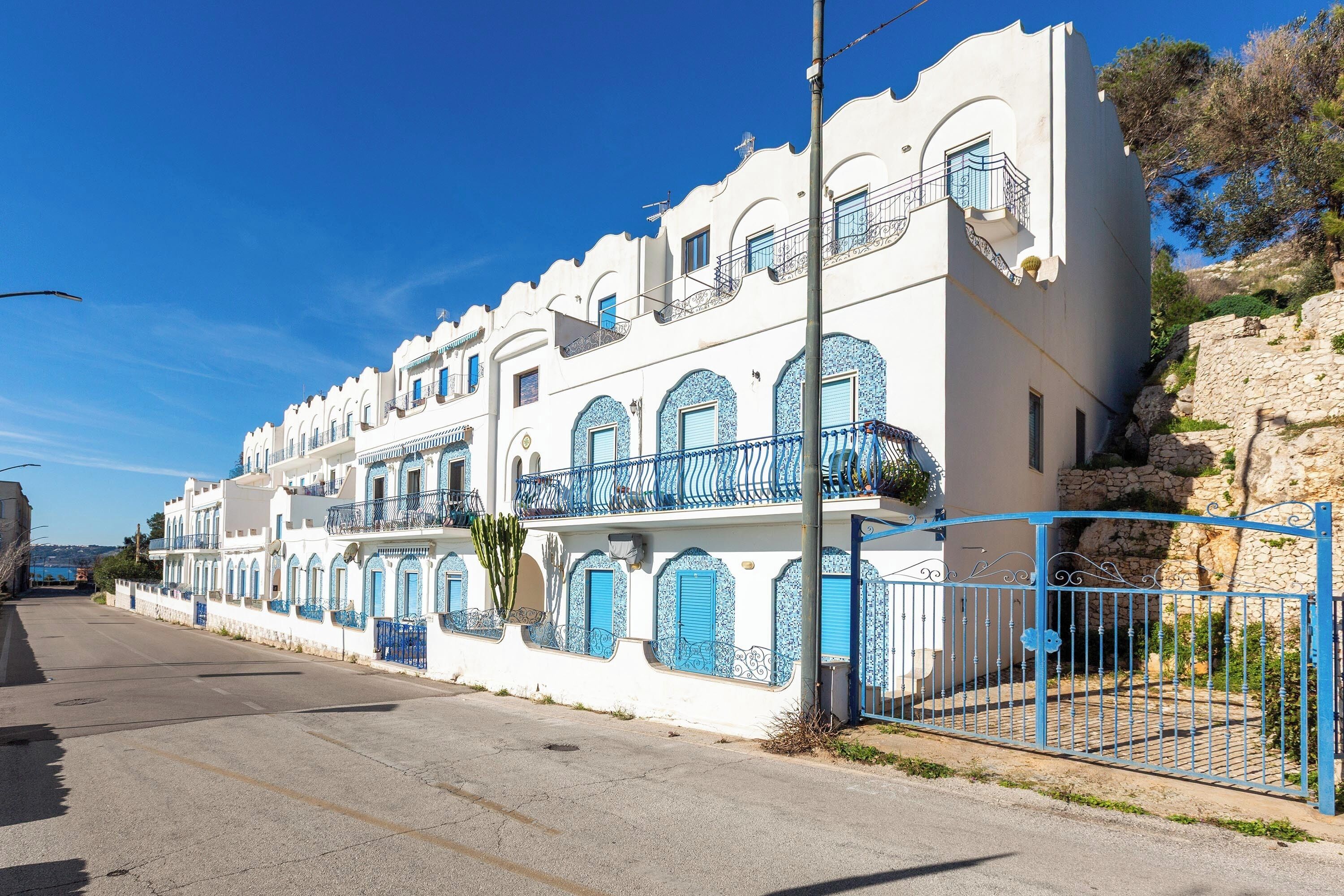 Une rangée de bâtiments blancs agrémentés de touches de bleu et dotés de balcons, un ciel dégagé et un mur de pierre sur la droite.