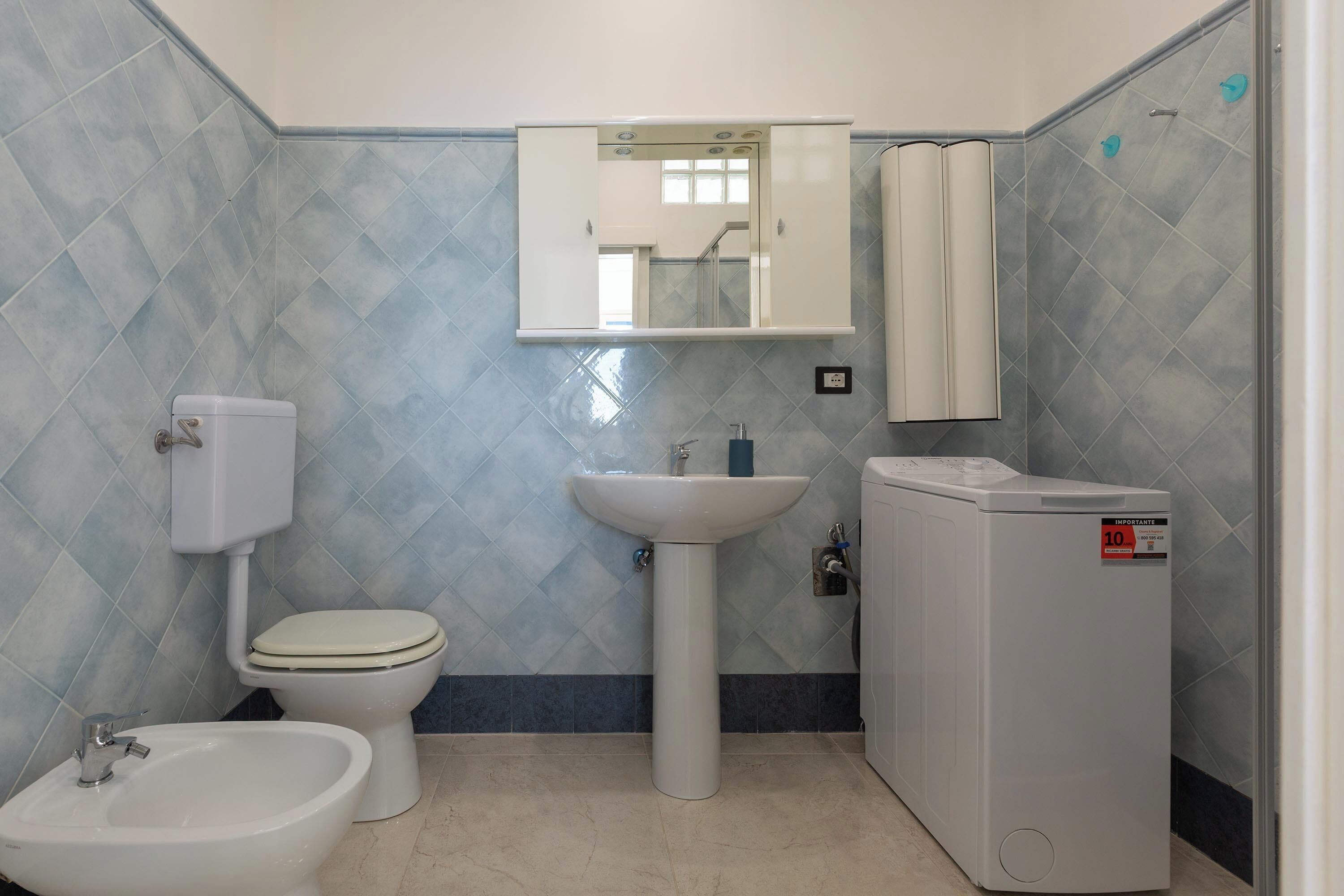 A bathroom with a toilet, bidet, sink, mirror, and a washing machine.