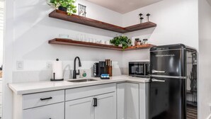 A modern kitchen with white cabinets, a black refrigerator, a microwave, and wooden shelves with plants. - Chic Yosemite Escape: King Suite w/ Plush Comforts (Oakhurst)