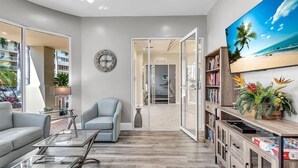 A modern living room with a glass coffee table, a sofa, a chair, a bookshelf, and a large TV screen displaying a beach scene. - Bahia Vista 363 (St. Petersburg)