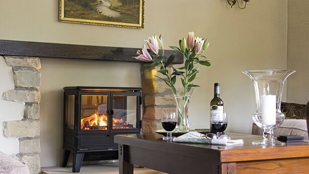 Una sala de estar acogedora con una chimenea de piedra, una mesa de madera con un jarrón de flores, copas de vino y una botella de vino.