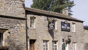 Un edificio de piedra con dos letreros que dicen "The Buck Inn". Hay mesas de picnic afuera.