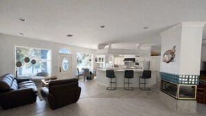 Amplia sala de estar con un sofá de cuero oscuro, una mesa de comedor y una cocina con isla central y taburetes de bar.