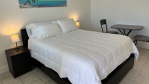 A neatly made bed with white bedding, a small bedside table with a lamp, a chair, and a painting of a beach scene on the wall.
