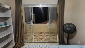 Et værelse med en skydedør af glas, der fører ud til en balkon med fliset gulv. Til venstre ses et hvidt reolsystem, og til højre står en ventilator.