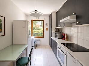 A modern kitchen with a dining area, a window with a view, and a clock on the wall. - Spacious 3-room apartment with a terrace – Quiet apartment complex with a pool (Mandelieu-la-Napoule)