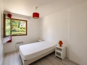 A minimalist bedroom with a single bed, a small bedside table, a nightstand, a window with a view of greenery, and a red pendant light. - Spacious 3-room apartment with a terrace – Quiet complex with a pool (Mandelieu-la-Napoule)