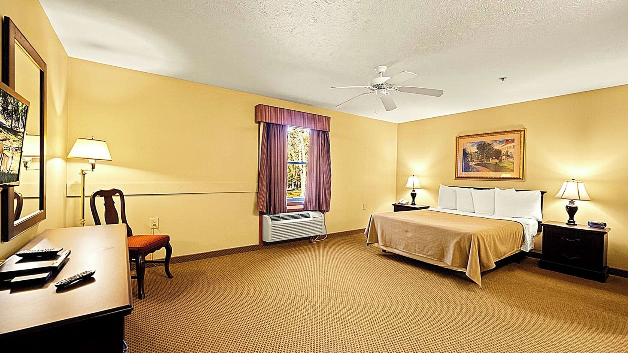 Une chambre d’hôtel comprenant un lit, des tables de chevet, un bureau, une chaise, un ventilateur de plafond et une fenêtre avec des rideaux.