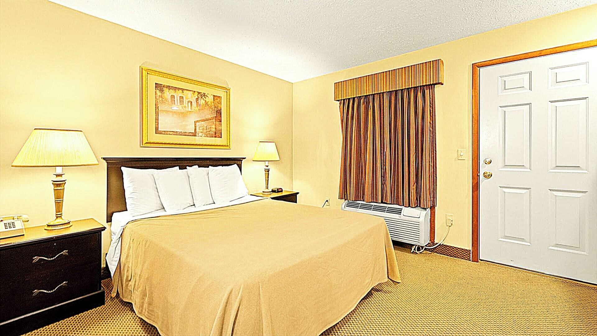 A hotel room with a bed, two bedside tables with lamps, a painting on the wall, a window with curtains, and a door.