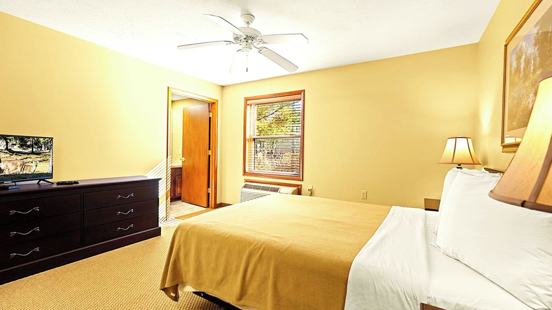 A hotel room with a bed, a dresser, a ceiling fan, a window with blinds, and a television on a dresser.
