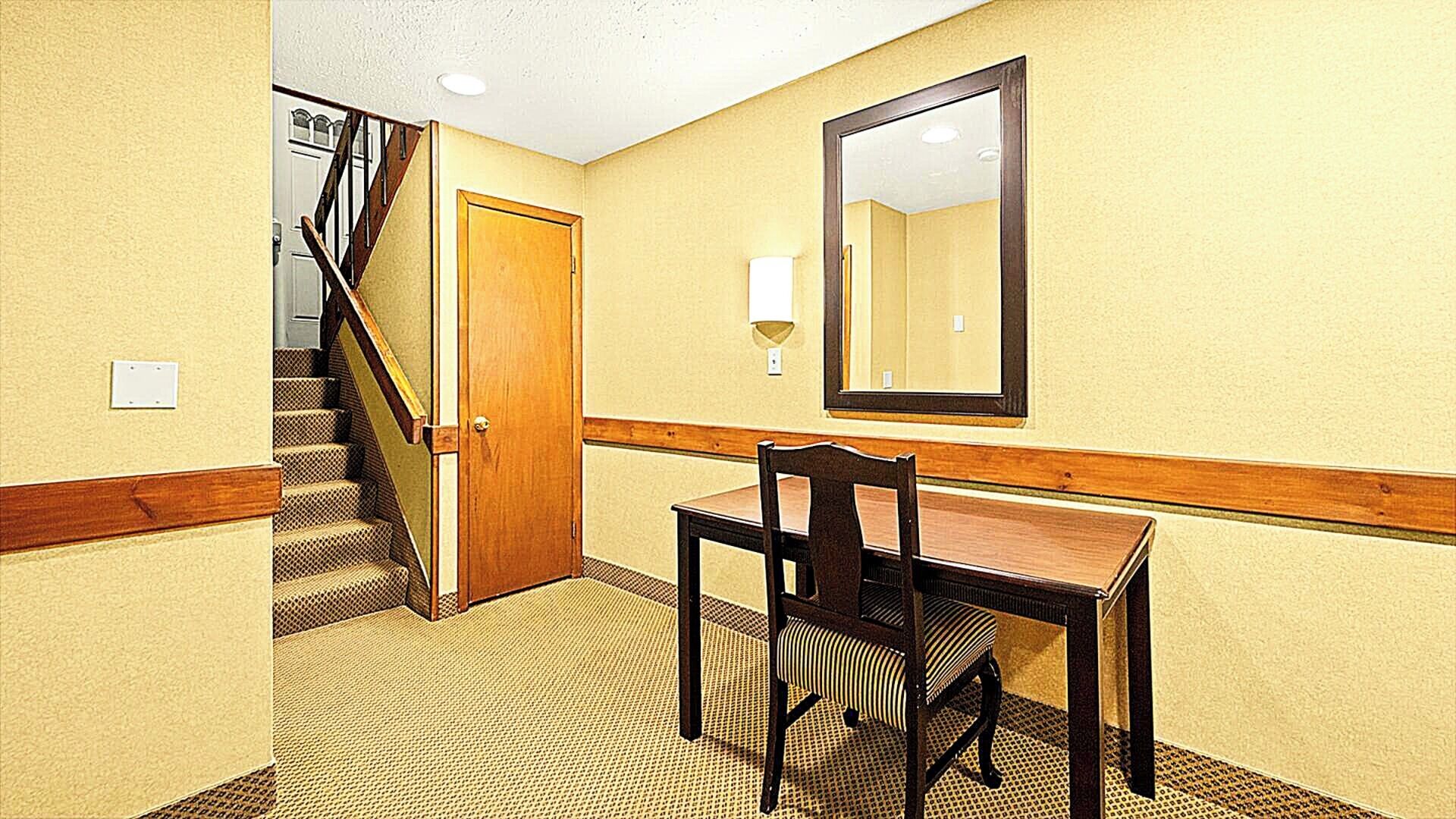 Habitación de hotel con escritorio de madera, silla y espejo en la pared. El suelo está alfombrado y hay una escalera que conduce a otra habitación.