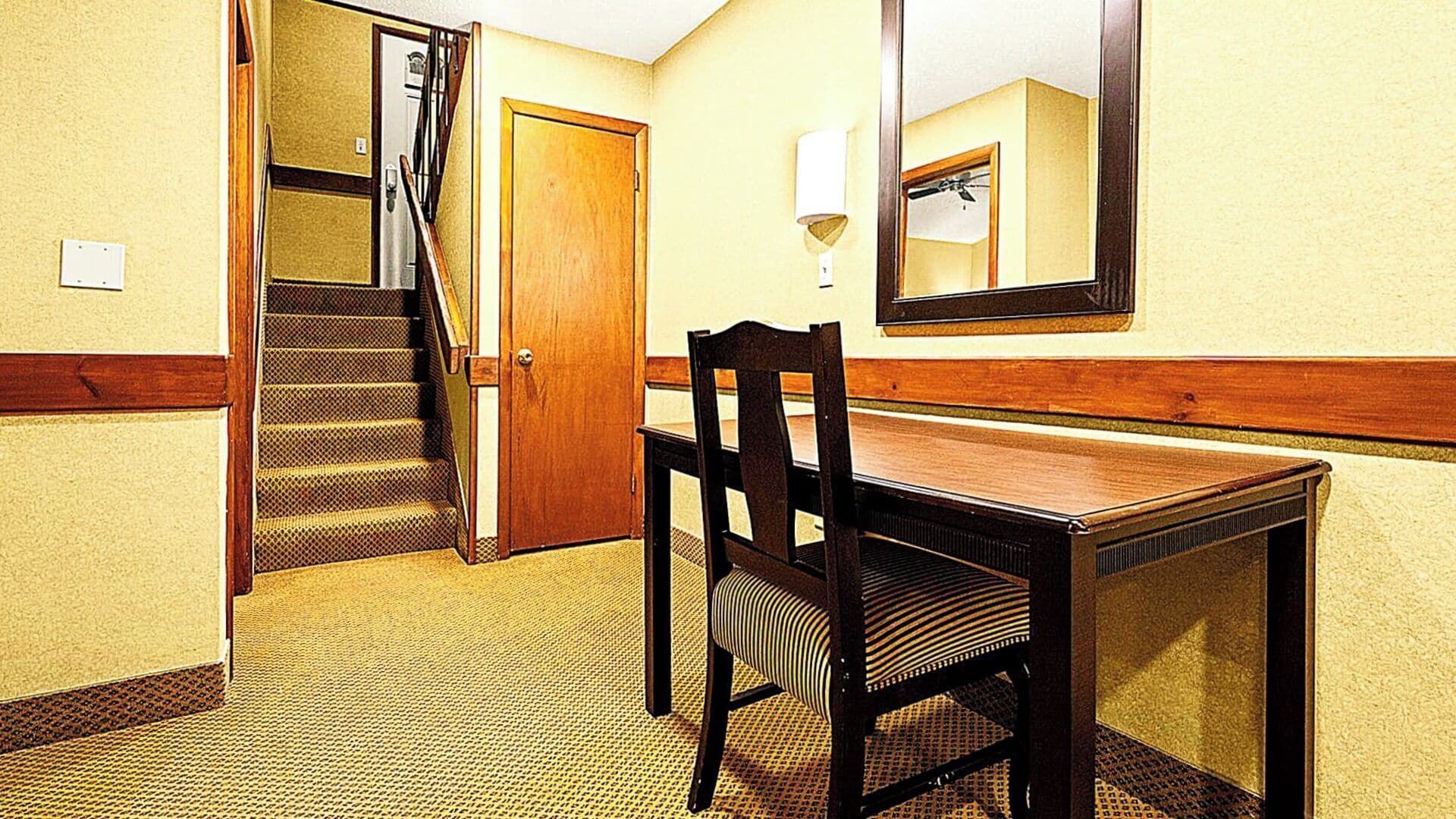 A hotel room with a wooden desk, a chair, a mirror, a staircase, and a bathroom visible through an open door.