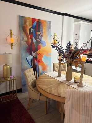 Un coin repas avec une table ronde en bois, des chaises recouvertes d’un tissu beige, un grand tableau abstrait au mur, un tapis rouge et un vase décoratif avec des bougies.