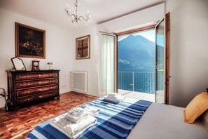 Ein Schlafzimmer mit Holzboden, einem Bett mit blau gestreifter Decke, einem Schminktisch, einer Uhr und einem Bild. Es gibt einen Balkon mit Blick auf das Meer und die Berge.