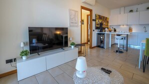 A modern living room with a large flat-screen TV, a white entertainment unit, a small table with a lamp, and a glass table with remote controls. - Residence degli Oleandri - APP. A1 (Argegno)