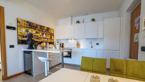 A modern kitchen with white cabinetry, a dining area with a white table and yellow chairs, and a large painting on the wall. - Residence degli Oleandri - APP. A1 (Argegno)